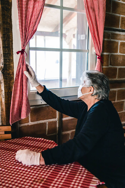 Elderly woman, grandma wearing a mask sitting inside the house by the window alone, looking through the window. Corona virus outbreak - Foto, imagen
