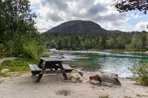 Camping área de descanso para piquenique com banco de mesa de madeira na areia pelo rio espumante frio em montanhas pinhal da Noruega
 - Foto, Imagem