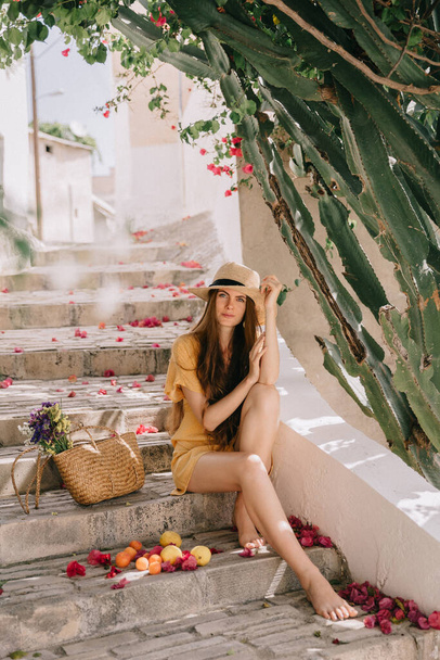 girl in a yellow dress and hat with long brown hair, is sitting on the steps, on the background of green flowering cactus and pink bougainvillea flowers, in summer in Mediterranean, on vacation - Foto, Imagen