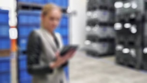 wazig blond vrouw dragen grijs uniform holding apparaat weg te kijken hurken in magazijn. - Foto, afbeelding