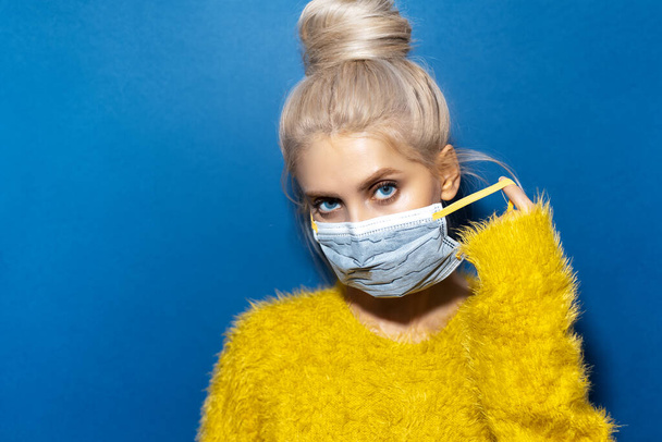 Studio portret młodej blondynki zdejmującej maskę medyczną przeciw koronawirusowi. Tło koloru niebieskiego. - Zdjęcie, obraz