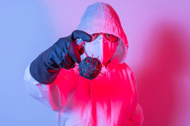 Man in beschermingspak, bril en masker spuitend alcohol, desinfecterend middel op een coronavirus model met blauwe en rode lichten - Foto, afbeelding