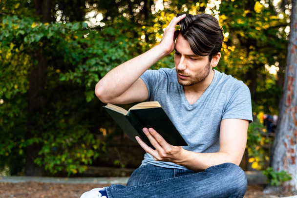 Człowiek czyta z zainteresowaniem starą książkę historyczną, siedzącą w parku w weekend. - Zdjęcie, obraz