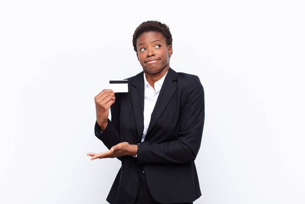 young pretty black womanshrugging, feeling confused and uncertain, doubting with arms crossed and puzzled look holding a credit card - Foto, imagen