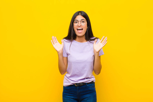 young pretty latin woman looking happy and excited, shocked with an unexpected surprise with both hands open next to face against flat wall - 写真・画像