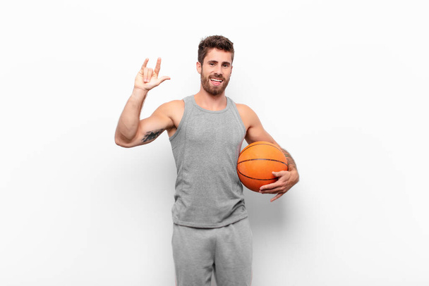 young handsome man feeling happy, fun, confident, positive and rebellious, making rock or heavy metal sign with hand holding a basketball ball. - Photo, image
