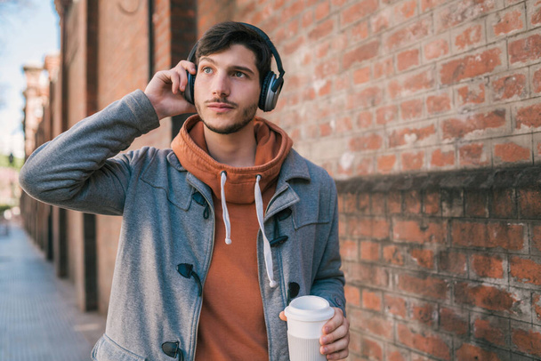 Portrait of young man with earphones and a cup of coffee against brick wall. Outdoors. Urban concept. - Photo, image