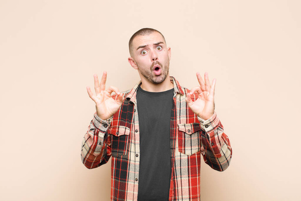 young handsome man feeling shocked, amazed and surprised, showing approval making okay sign with both hands against flat wall - Fotoğraf, Görsel