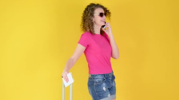 Attractive young girl in sunglasses with a suitcase and a ticket is using a smartphone isolated over yellow orange background in studio. journey concept. - Filmagem, Vídeo