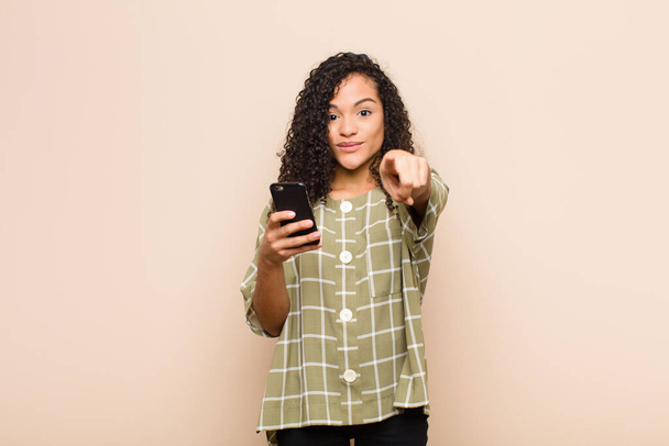 young black woman pointing at camera with a satisfied, confident, friendly smile, choosing you with a smartphone - Photo, image