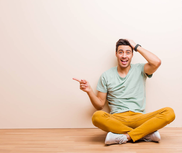 young arabian man laughing, looking happy, positive and surprised, realizing a great idea pointing to lateral copy space sitting on the floor - Photo, Image