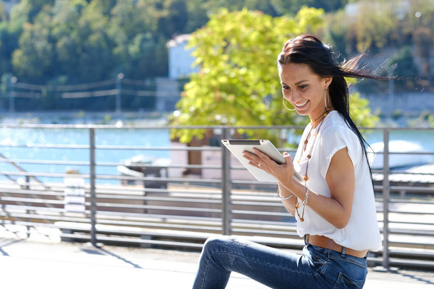 Giovane bella ragazza capelli neri nel vento utilizzando tablet al di fuori in estate per strada con sfondo verde fiume
 - Foto, immagini