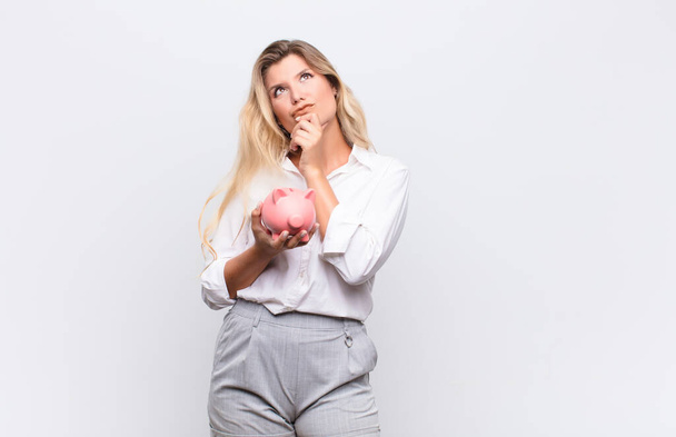 young pretty latin woman thinking, feeling doubtful and confused, with different options, wondering which decision to make with a piggy bank against white wall - Photo, Image