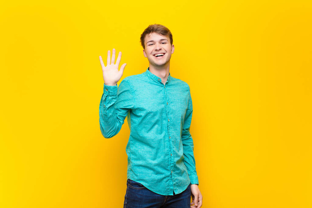 young blonde man smiling happily and cheerfully, waving hand, welcoming and greeting you, or saying goodbye isolated against flat wall - Photo, image
