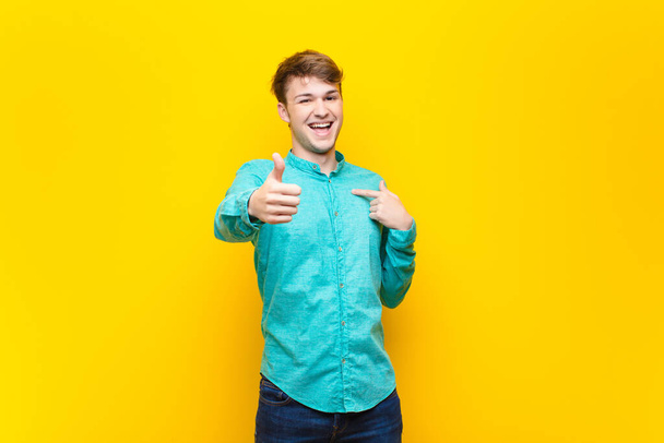 young blonde man feeling proud, carefree, confident and happy, smiling positively with thumbs up isolated against flat wall - Fotoğraf, Görsel