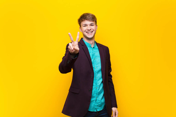 young blonde man smiling and looking friendly, showing number two or second with hand forward, counting down isolated against flat wall - Fotoğraf, Görsel