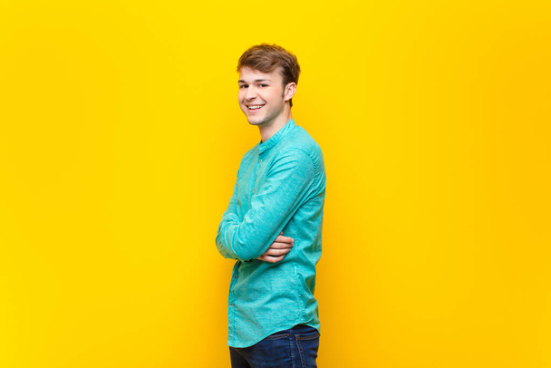young blonde man smiling to camera with crossed arms and a happy, confident, satisfied expression, lateral view isolated against flat wall - Фото, зображення
