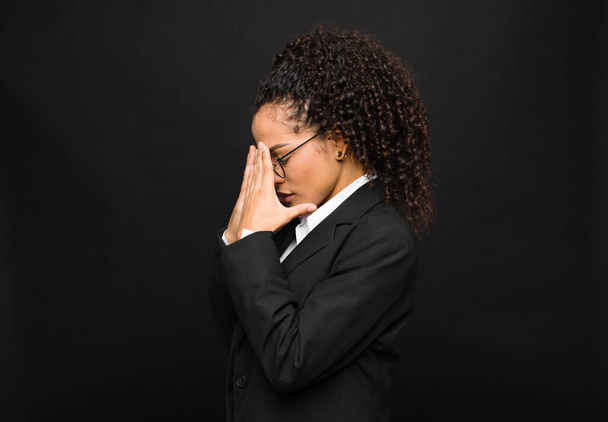 jonge zwarte vrouw bedekken ogen met handen met een trieste, gefrustreerde blik van wanhoop, huilen, zijaanzicht tegen zwarte muur - Foto, afbeelding