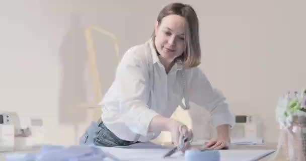 Slow motion video of the fashion designer works at a table with curves and a pattern, she are cutting fabric, around lies scissors, centimeter, sew machine and dummy on the background, Smooth movement - Filmati, video