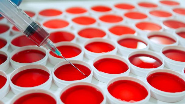 Detail injekční stříkačky odebírající vzorek červené tekutiny ze skupiny kulatých červených klinických vzorků na bílém povrchu - Fotografie, Obrázek