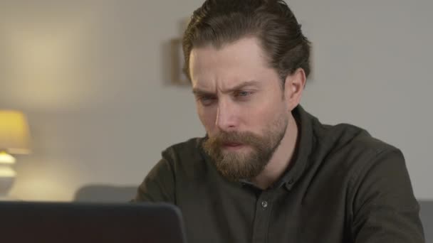 freelancer met een baard zit aan een tafel in de kamer, kijkt naar een laptop monitor, een man is moe, hij gaapt. - Video