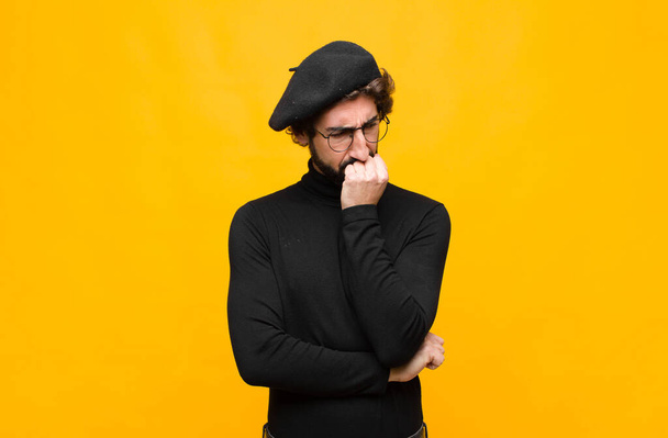 young french artist man feeling serious, thoughtful and concerned, staring sideways with hand pressed against chin against orange wall - 写真・画像