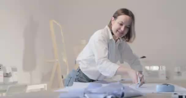 Slow motion video of the fashion designer works at a table with curves and a pattern, she are cutting fabric, around lies scissors, centimeter, sew machine and dummy on the background, Smooth movement - Felvétel, videó