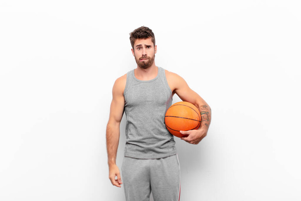 young handsome man feeling clueless, confused and uncertain about which option to pick, trying to solve the problem holding a basketball ball. - 写真・画像