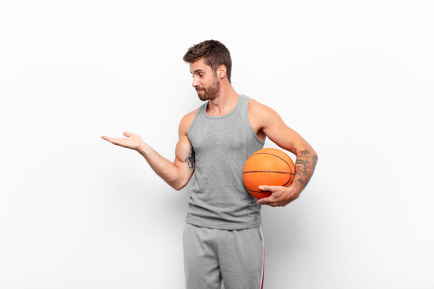 young handsome man feeling happy and smiling casually, looking to an object or concept held on the hand on the side holding a basketball ball. - Фото, зображення