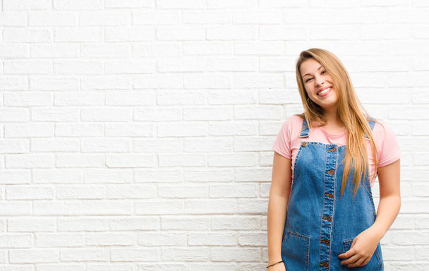 young blonde woman smiling cheerfully and casually with a positive, happy, confident and relaxed expression against brick wall - Foto, afbeelding