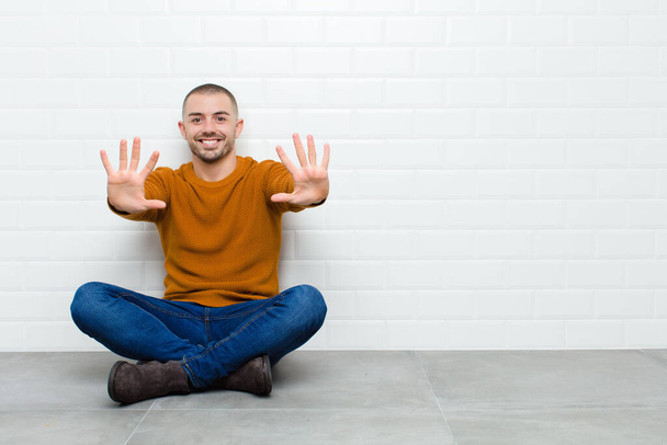 young handsome man smiling and looking friendly, showing number ten or tenth with hand forward, counting down sitting on the floor - Foto, afbeelding