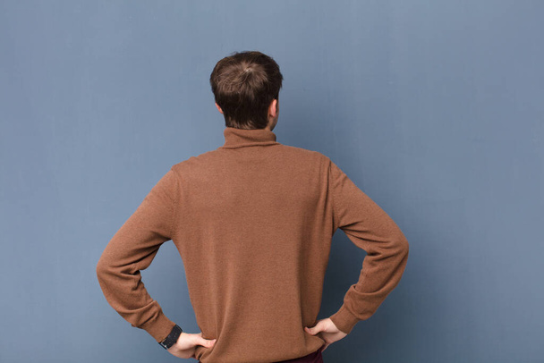 young blonde man feeling confused or full or doubts and questions, wondering, with hands on hips, rear view isolated against flat wall - Foto, Bild