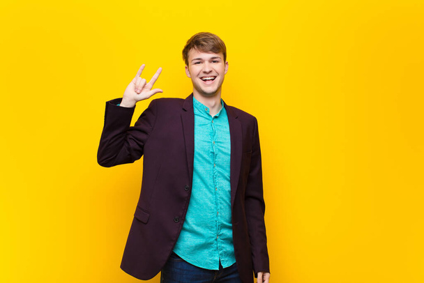 young blonde man feeling happy, fun, confident, positive and rebellious, making rock or heavy metal sign with hand isolated against flat wall - Photo, image