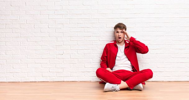young blonde man looking happy, astonished and surprised, smiling and realizing amazing and incredible good news sitting on the floor - 写真・画像
