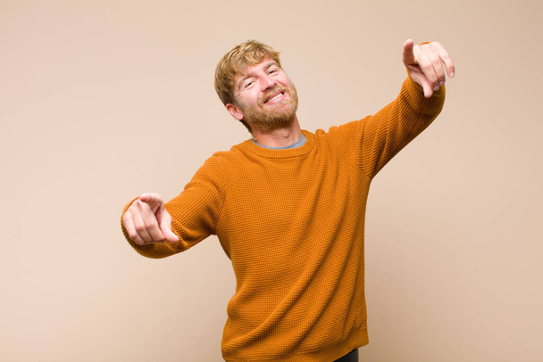 young blonde man feeling happy and confident, pointing to camera with both hands and laughing, choosing you against flat wall - Foto, Imagem