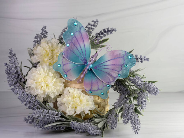 Corona de lavanda púrpura con claveles blancos y una gran mariposa de malla azul y púrpura decorada con piedras preciosas sentadas sobre un fondo de arenisca. Bonita decoración del hogar de primavera
. - Foto, Imagen