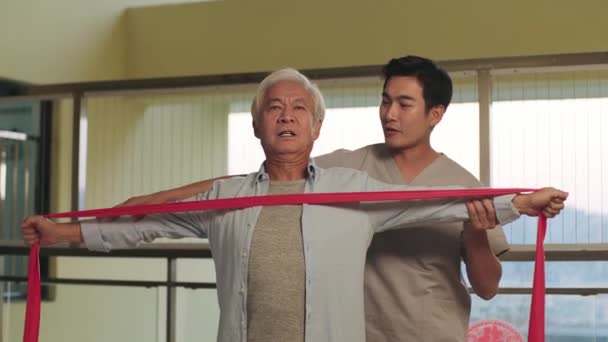 senior asian man rehabilitating using resistance band with help from physical therapist - Footage, Video
