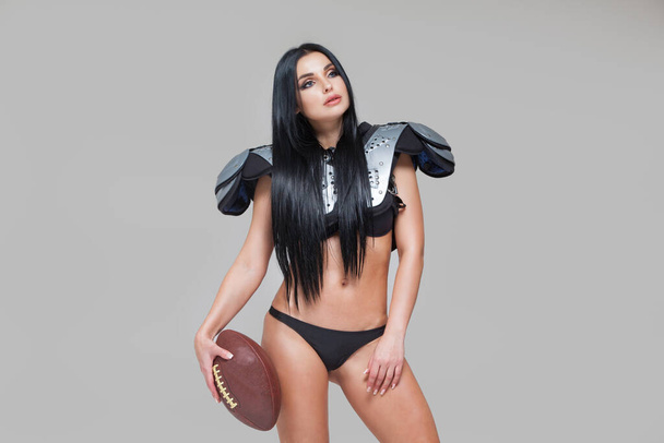 Beautiful sexy brunette female american football player in uniform posing with a ball isolated on grey background - Photo, Image