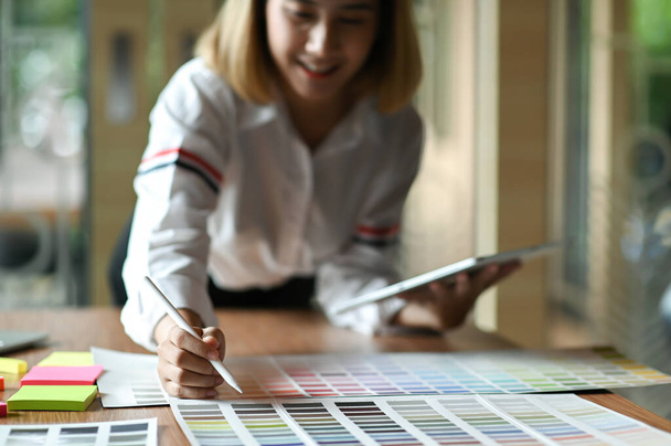Asian female graphic designer working in office. She uses tablet and point the pen on color chart. - Foto, immagini