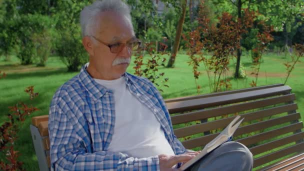 Starší muž sedící na lavičce a čtoucí v parku legrační knihu. - Záběry, video