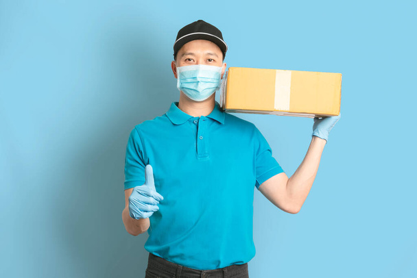 青いシャツの制服顔マスク手袋の配達人の従業員は青い背景に空の段ボール箱を保持します。コロナウイルス2019のコンセプト - 写真・画像