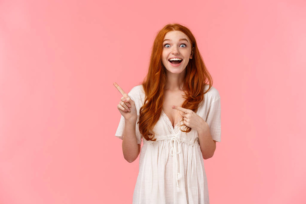 Excited pretty caucasian redhead girl in lovely spring white dress, pointing fingers left and smiling astonished, left speechless after visiting awesome event, left with positive impression - Photo, image