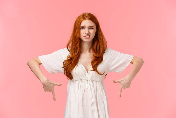 Well I dont know. Waist-up portrait indecisive and unsure, slightly displeased cute redhead woman in white dress, pointing down, cringe as express dislike, disappointed with gift, pink background - Фото, изображение