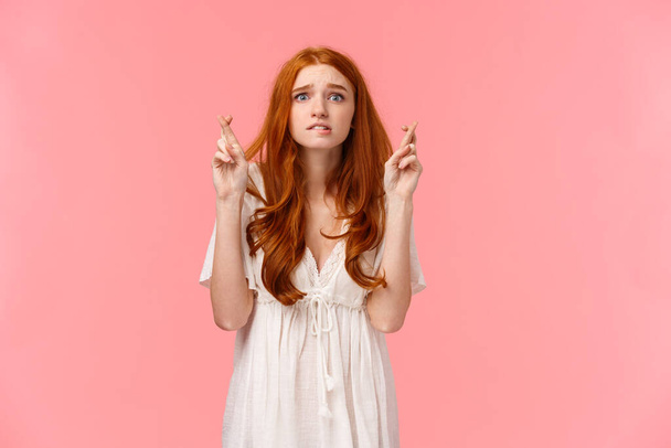 Nervous cute teenage redhead girl praying someting good happen, wish enter cool unversity, waiting enrollment info, cross fingers good luck, biting lip anxious, standing pink background - Photo, Image