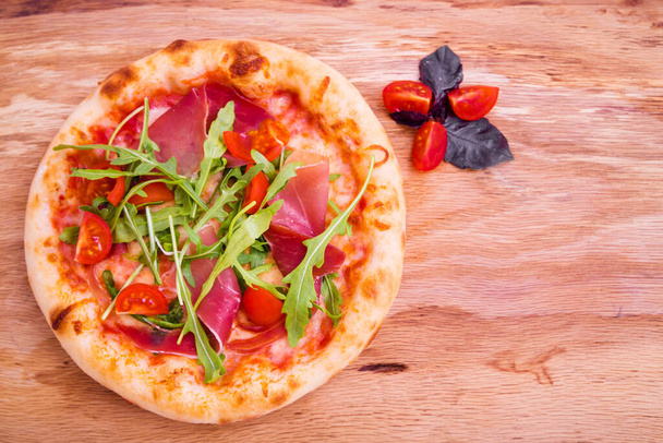Pizza with ham, tomatoes, arugula on a wooden background. Basil leaves and tomato slices as a decoration. Horizontal photo - Photo, Image