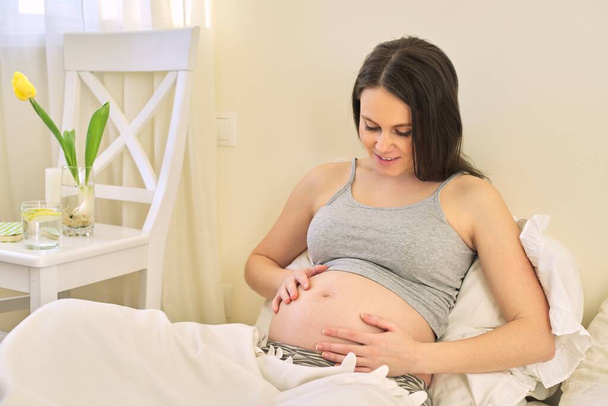 Young beautiful pregnant woman sitting at home in bed, female touching her tummy, waiting for the birth of baby - Foto, Imagem