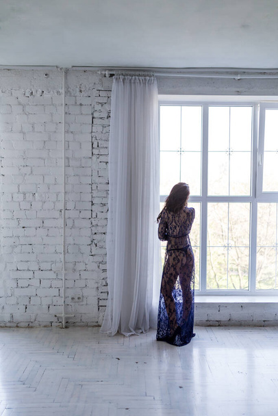 сексуальна дівчина в блакитному пігирі дивиться у вікно. простора біла кімната з величезним вікном. сонячний день красива брюнетка
 - Фото, зображення