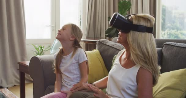 Caucasian girl enjoying her time at home, sitting on a couch with her grandmother, a woman is wearing a vr headset, social distancing and self isolation in quarantine lockdown during coronavirus covid19 epidemic, in slow motion - Materiaali, video