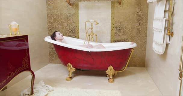 Blick auf eine junge kaukasische Frau in einem modernen Badezimmer, entspannen in einem Schaumstoff mit einem Gesichtspaket auf Badewanne zu Hause oder Hotelbad lächelnd verspielt genießen Luxus-Lifestyle.Black magic Kamera. 4k - Filmmaterial, Video