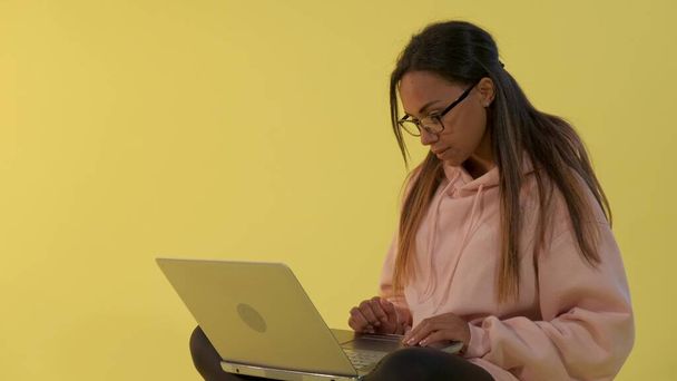 黄色の背景にコンピュータ上で働く混合レースの若い女性 - 写真・画像
